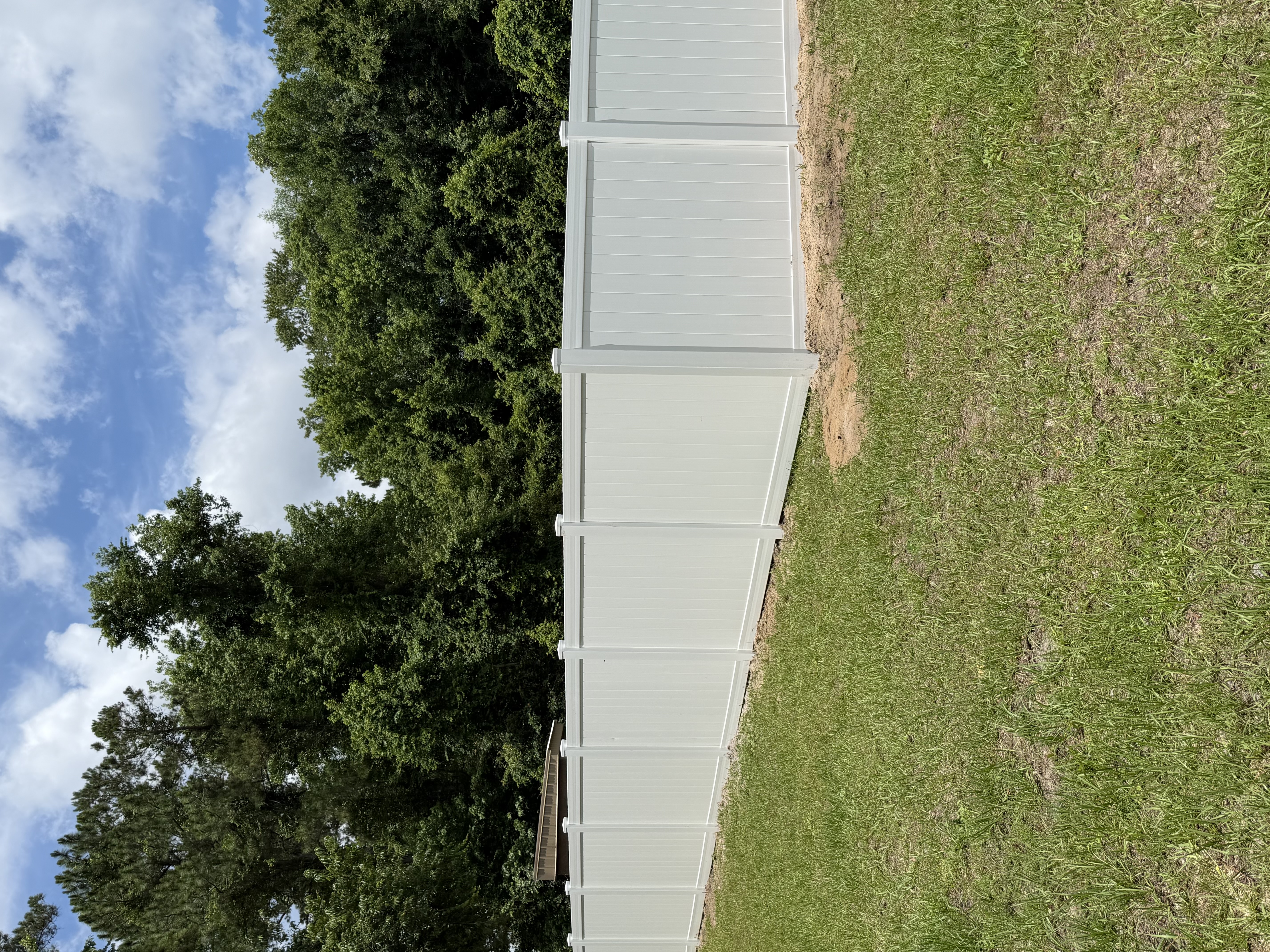 White vinyl privacy fence running along the edge of a grass yard with trees behind it